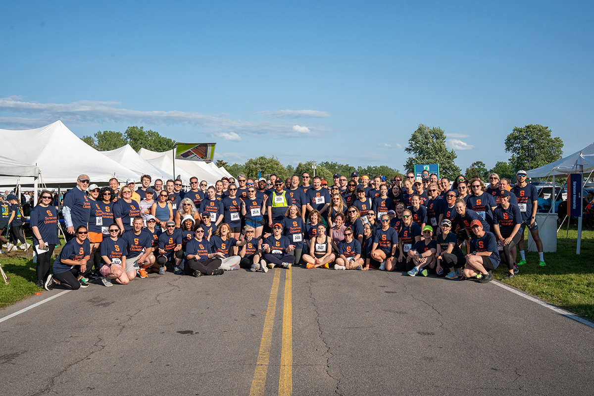 A large group of people pose for a photo on a road, wearing matching navy blue shirts with race bibs at the Syracuse WorkForce Run. White tents are visible in the background under a clear, sunny sky.