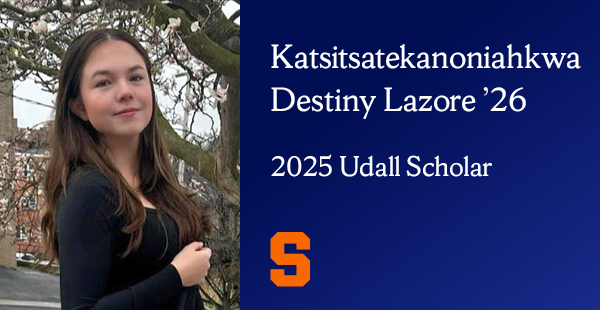 A blue gradient graphic features a person standing outdoors in front of a tree with blooming flowers. The person is wearing a black long-sleeve top and blue jeans, holding a black bag with floral embroidery. The background includes a building and a road. The graphic also includes the text "Katsitsatekanoniahkwa Destiny Lazore ’26, 2025 Udall Scholar" and includes an orange Block S.