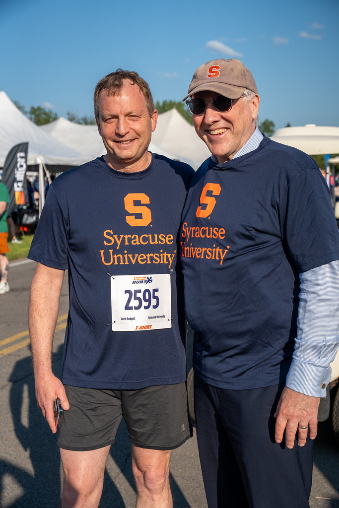 Two individuals stand side by side outdoors, both wearing navy blue t-shirts with the orange Syracuse University logo and text. One person has a race bib numbered 2595. A tent and other people are visible in the background.