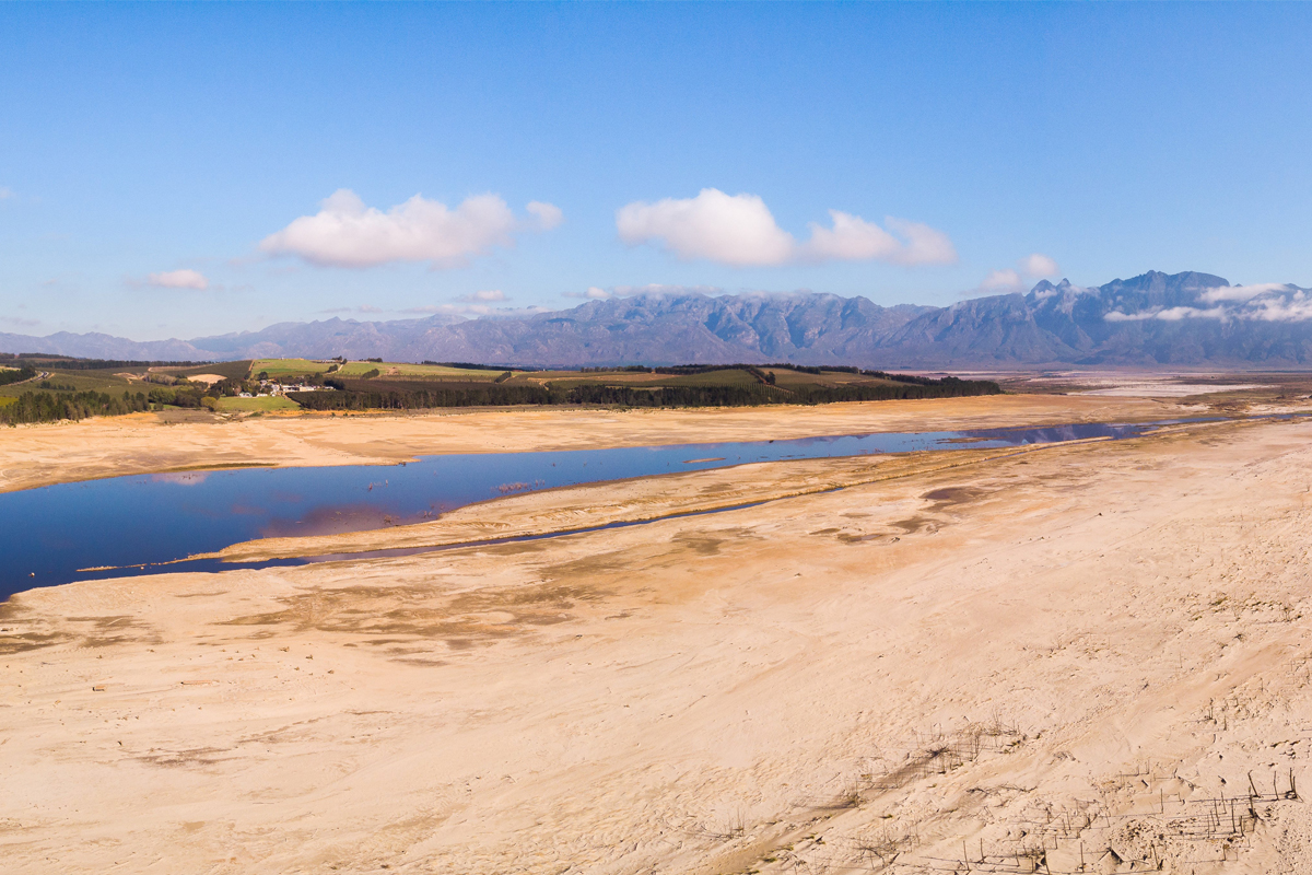 Expansive arid landscape with a narrow stream or body of water cutting through the center. The terrain is dry and barren, featuring patches of cracked soil and sparse vegetation. Mountains rise in the background beneath a mostly clear blue sky with a few scattered clouds.