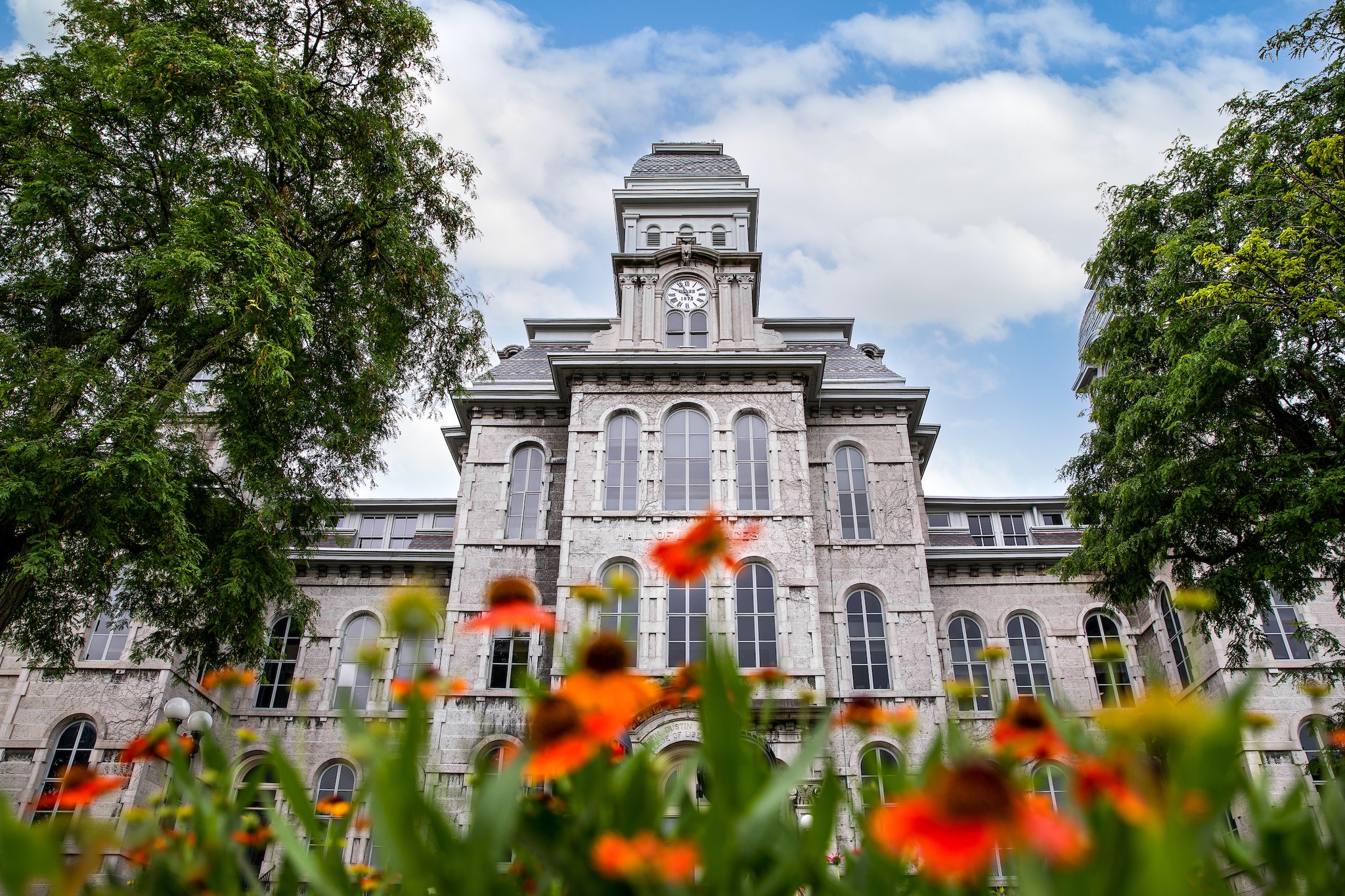 A view of the Hall of Languages at Syracuse University, featuring its central clock tower and arched windows. The building is framed by green trees on both sides, with orange flowers in the foreground against a partly cloudy blue sky.