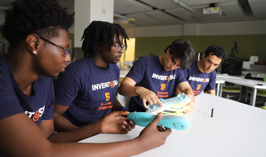 Four individuals in 'INVENT@SU' Syracuse University shirts sit at a classroom table, examining a pair of athletic shoes.