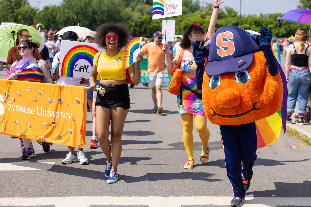 Group of people in a parade, including Syracuse University mascot Otto the Orange, holding rainbow flags and signs. One sign reads 'GAY' and another person carries an orange 'Syracuse University' banner. Participants wear bright clothing and accessories