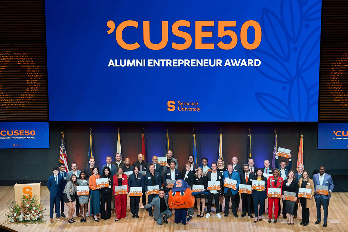 A group of people stands on a stage holding signs that read 'Syracuse University 'CUSE50 Alumni Entrepreneur Award.' Behind them is a large blue screen displaying the same text along with the Syracuse University logo. The stage features flags in the background and a podium with flowers on the left side.