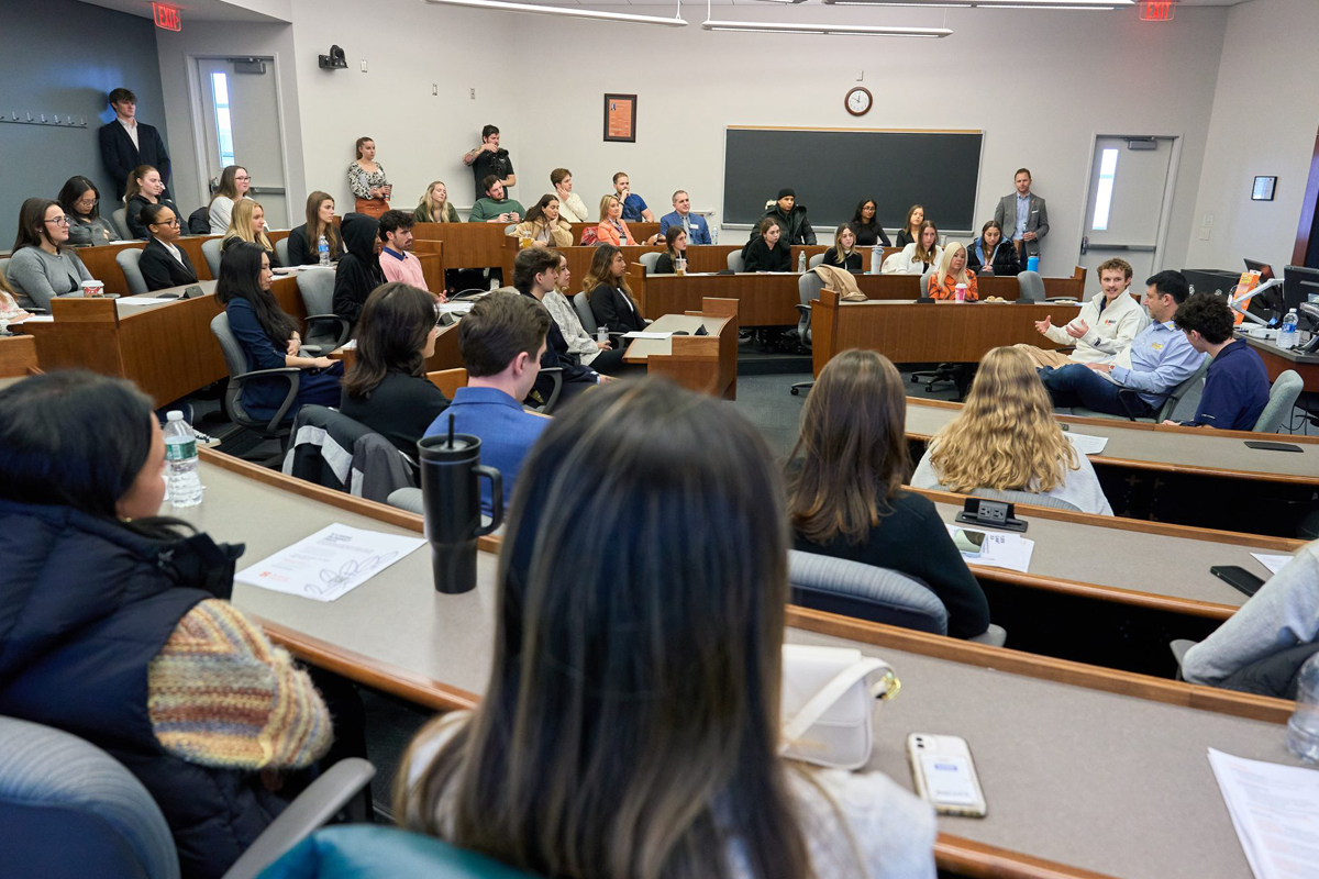 A well-lit classroom with students seated in a semi-circular arrangement at wooden desks, facing a blackboard and a clock at the front. Some students are taking notes, while others listen to a person standing near the blackboard.