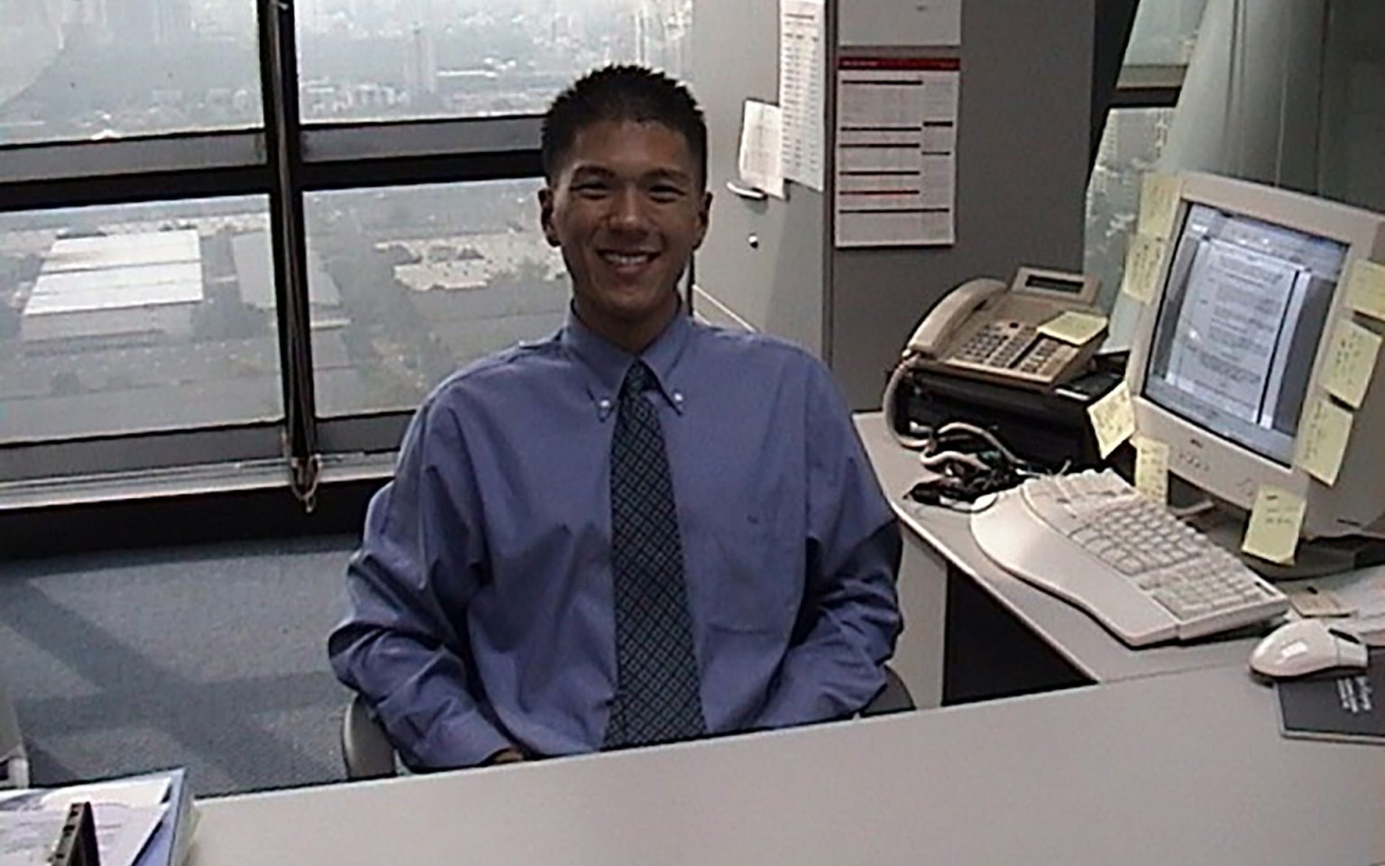 A person in a blue dress shirt and patterned tie sits at a desk in an office. The desk holds a computer monitor with text, sticky notes, a telephone, and office supplies. Large windows behind them reveal an urban landscape with buildings and roads.