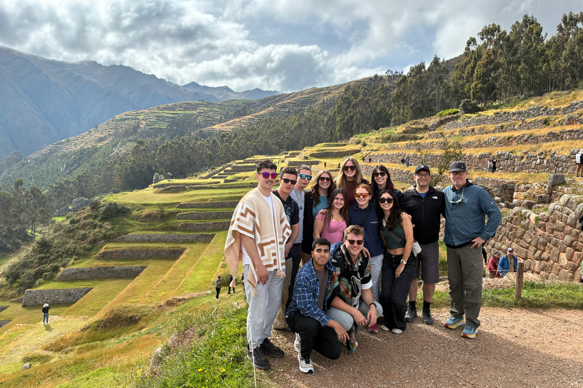 A group of people poses in front of the ancient Inca terraces at Moray, Peru. The background showcases circular terraced agricultural fields carved into a green hillside, with mountains and a partly cloudy sky in the distance. The group is casually dressed.