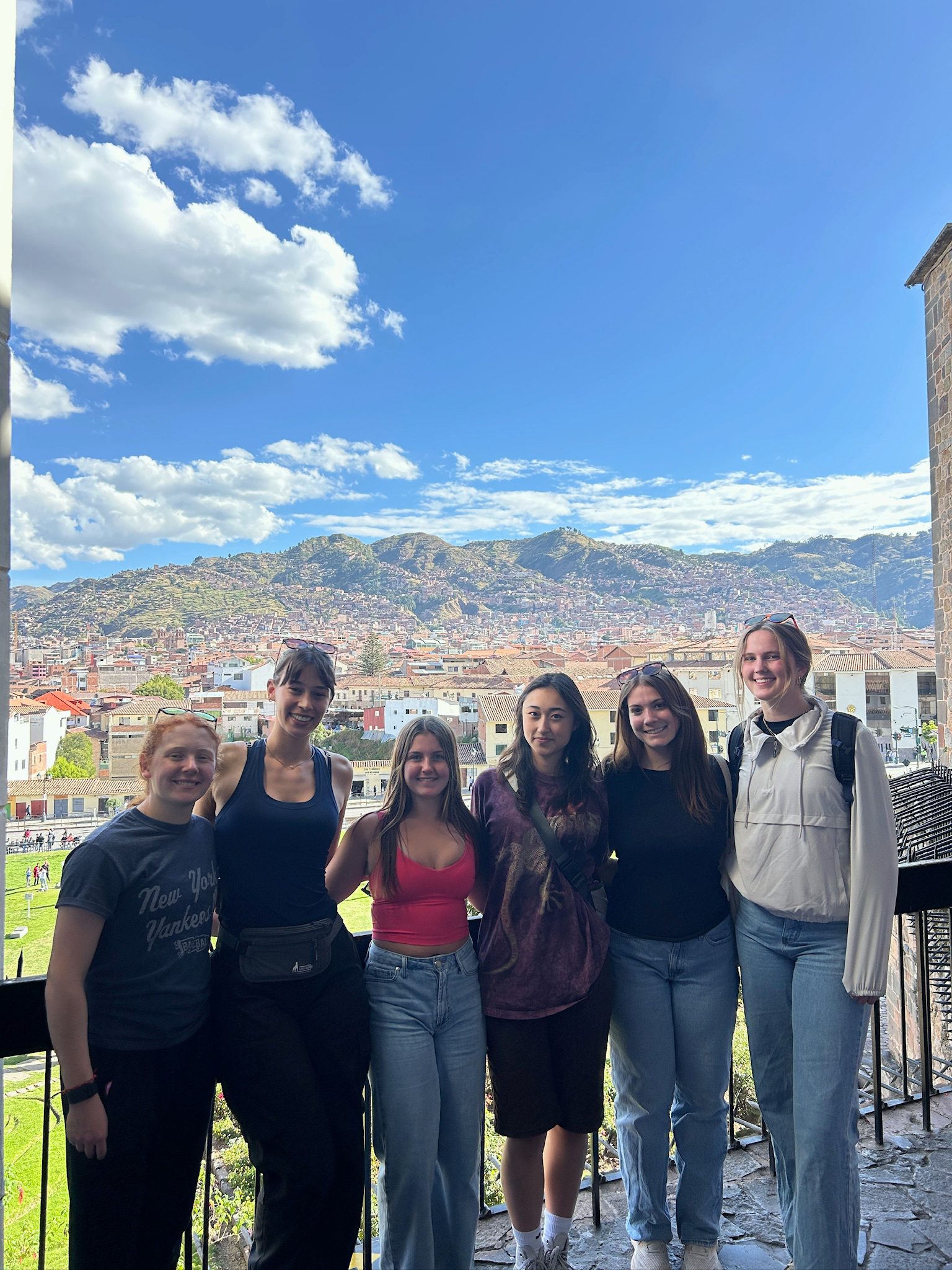 A group of six people stands on a balcony overlooking a scenic cityscape with red-roofed buildings and lush green mountains in the background. The sky is mostly clear with a few clouds. 