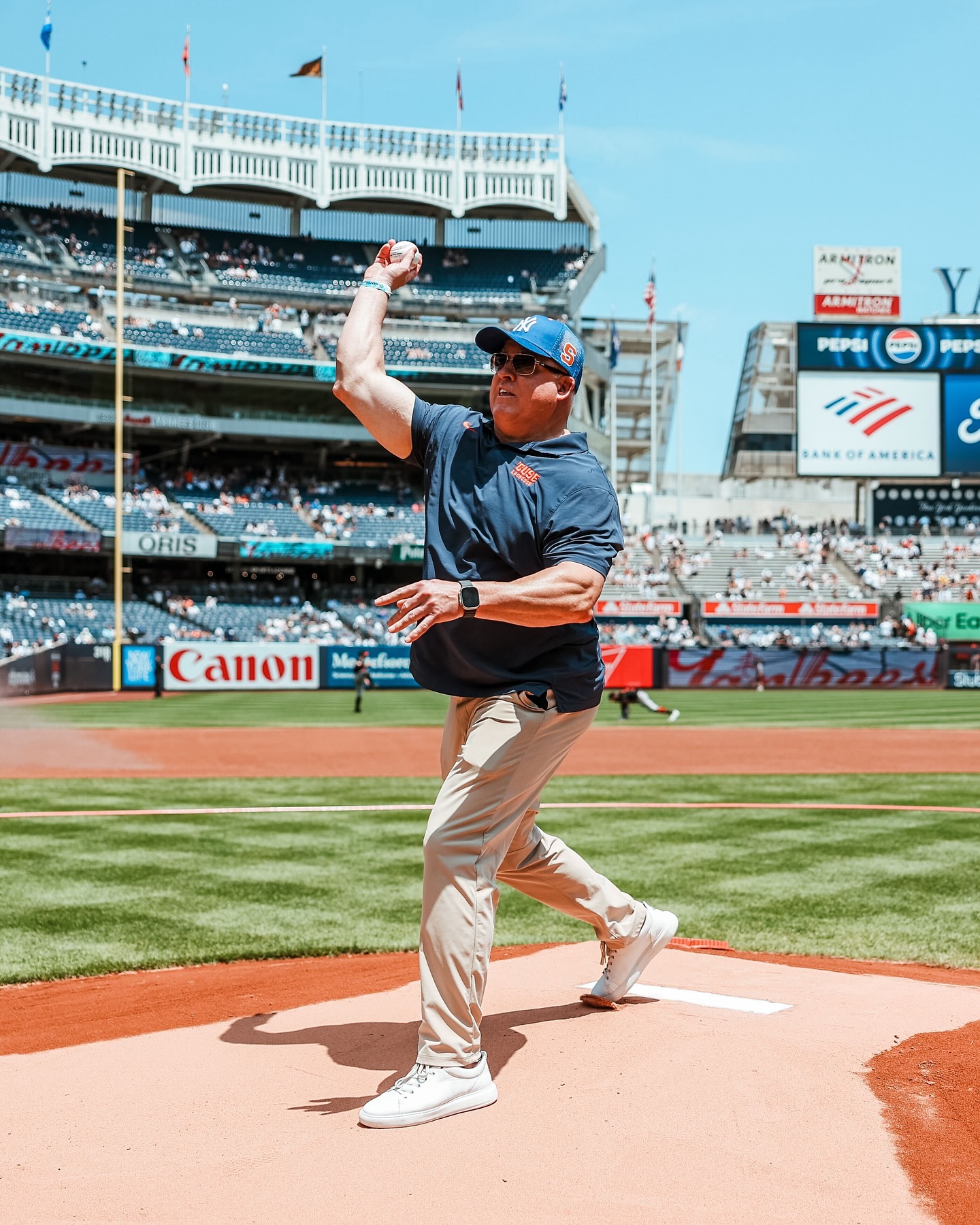 A person stands on a pitcher's mound in a baseball stadium, poised to throw a baseball. The stadium is filled with spectators, and colorful advertisements line the outfield walls. The individual is dressed in a navy blue shirt, beige pants, white sneakers, and a watch. 