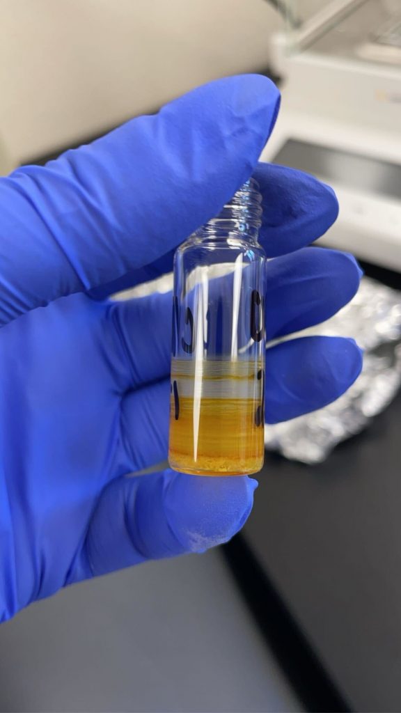 A gloved hand holding a small, clear glass vial containing a liquid with two distinct layers: a yellowish-orange layer at the bottom and a clear layer on top. The background is a blurred laboratory setting with visible equipment.