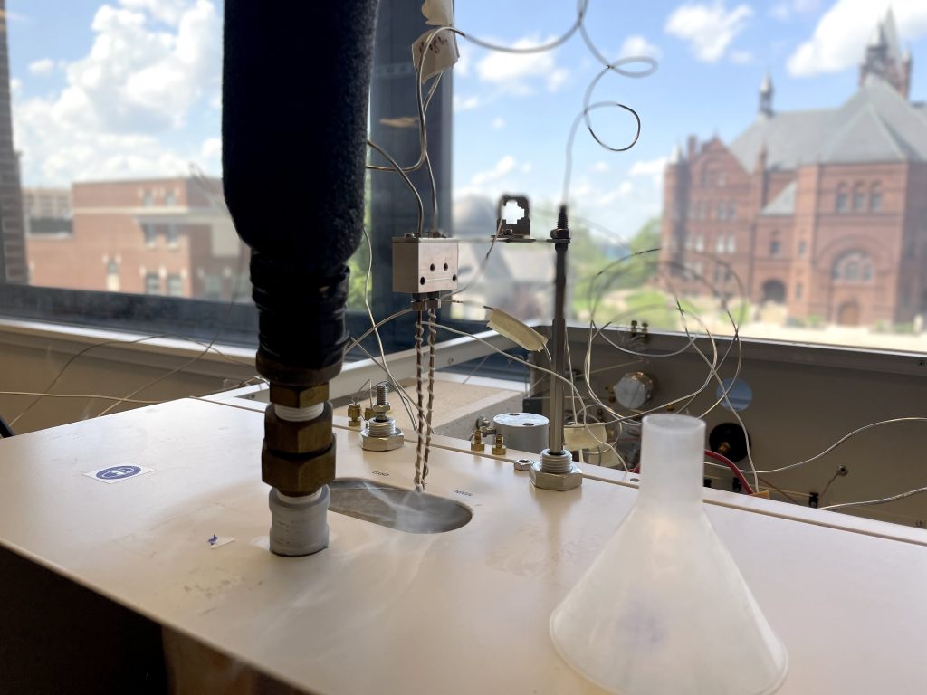 Laboratory setup with scientific equipment on a table, including a white funnel, tubes, and wires connected to various apparatuses. In the background, a large window reveals a view of buildings, one with red brick architecture, under a partly cloudy blue sky.