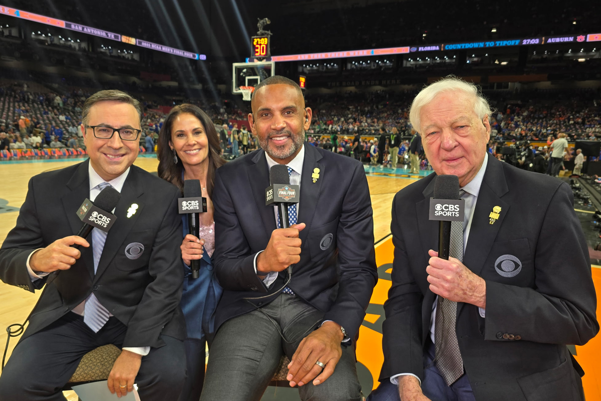 Four individuals sit courtside at a basketball game, holding CBS Sports-branded microphones. They are dressed in formal attire, including suits and ties. Behind them, players are warming up on the court, and spectators fill the stands.