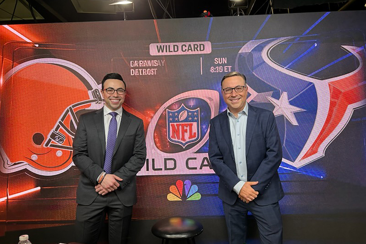 Two individuals stand in front of a large screen displaying promotional graphics for an NFL Wild Card game. The screen shows the logos of the Cleveland Browns and the Houston Texans, with text reading "WILD CARD," "CLEVELAND AT HOUSTON," "SUN 8:15 ET," and includes the NFL and NBC logos.