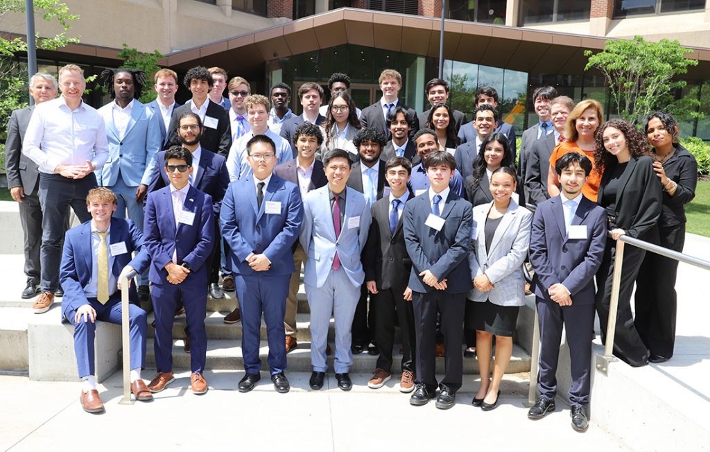 Invent@SU participants, judges and audience members pose on steps outside a building with large windows and greenery.