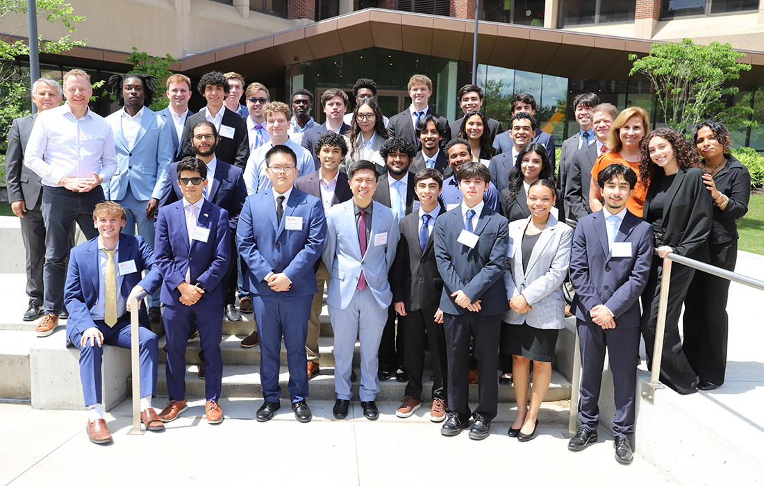Invent@SU participants, judges and audience members pose on steps outside a building with large windows and greenery.