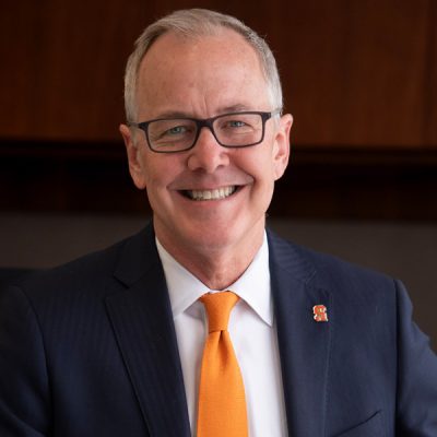 A person wearing a dark suit, white shirt, and orange tie. There is an orange Block S pin on the left lapel of the suit jacket.