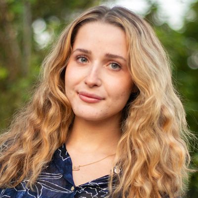 A person with long, wavy blonde hair is shown from the shoulders up. The person is wearing a dark blue shirt with a white floral pattern and a delicate gold necklace. The background consists of greenery, suggesting an outdoor setting.