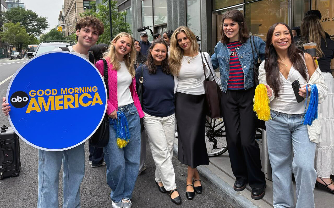 A group of six people stands on a sidewalk in front of a building. One person on the left holds a large circular sign that reads "ABC GOOD MORNING AMERICA" in yellow and white text on a blue background. Two others hold blue and yellow pom-poms. The group poses for a photo, with some smiling and looking at the camera. An urban street with buildings, trees, and pedestrians is visible in the background.