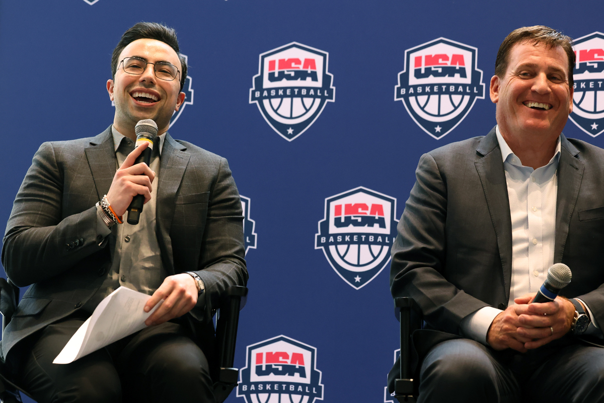 Two individuals sit in front of a backdrop featuring repeated USA Basketball logos. Both are dressed in suits and holding microphones; the person on the left also holds some papers.