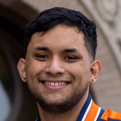 A person with short, dark hair and earrings is shown. The background appears to be an architectural structure with a curved arch and detailed stonework. The person is wearing an orange and blue jacket.