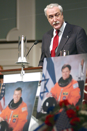 A man in a suit and red tie stands at a podium with a microphone, delivering a speech. In front of the podium are three large photographs of astronauts in orange spacesuits with their faces obscured. The background features the Israeli and American flags, with floral arrangements at the base of the display.