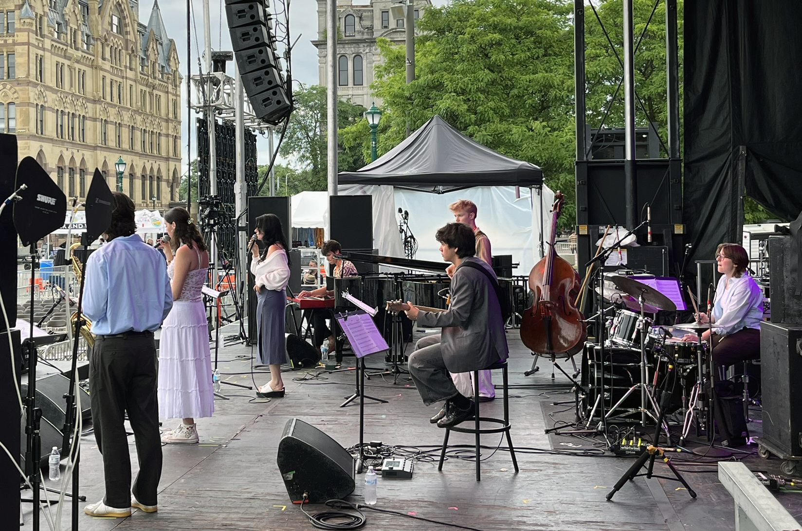 A group of musicians performs on an outdoor stage. They play instruments including a trumpet, keyboard, guitar, double bass, and drums. Some are standing, others are seated. The background features large, ornate buildings and green trees. Stage equipment like speakers and microphones is also visible.