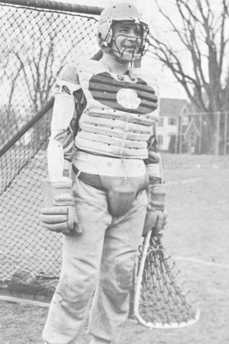 Black and white photo of a person in gear—helmet, chest protector, and shin guards—standing in front of a chain-link fence with leafless trees in the background. 