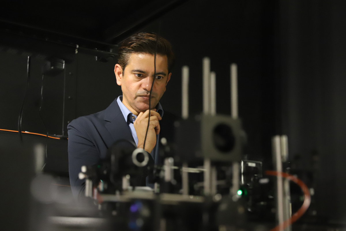 A person in costume is held behind a complex arrangement of scientific or technical equipment on a table. The configuration includes metal stems, lenses and electronic components, suggesting a laboratory or a research environment.