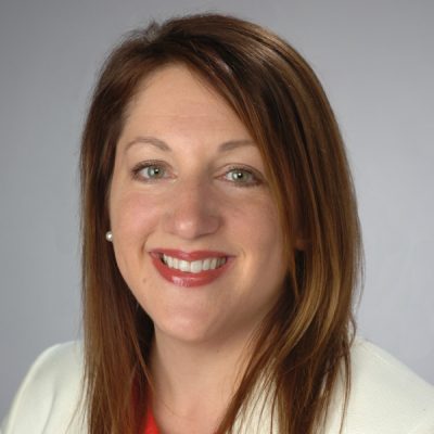 A person with shoulder-length brown hair wearing a white blazer over a red top. The background is plain and light gray.