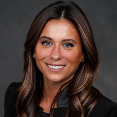 A professional headshot of a person with long, wavy hair wearing a black blazer. The background is a plain dark gray.