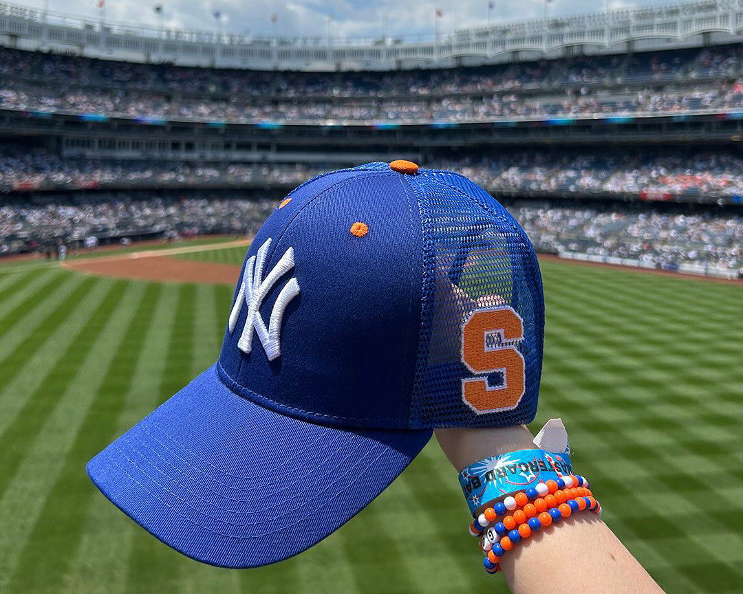 Person holding a blue New York Yankees cap with an orange Block S on the side, in a baseball stadium filled with spectators. The person is wearing colorful bracelets.