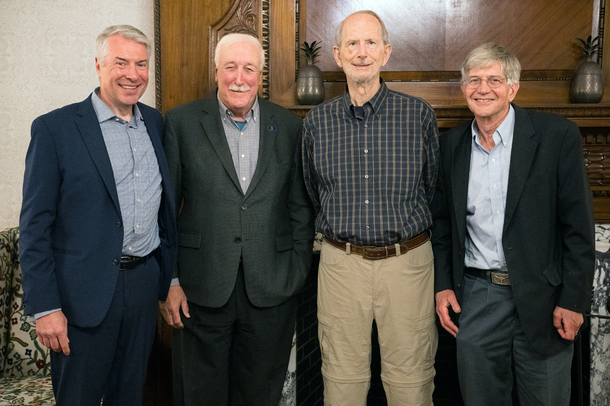 Four men stand side by side in front of a wooden wall with decorative elements. From left to right: the first man wears a blue suit with a light blue shirt; the second wears a dark suit with a checkered shirt; the third is dressed in a checkered shirt and beige pants; and the fourth wears a dark blazer over a light blue shirt.