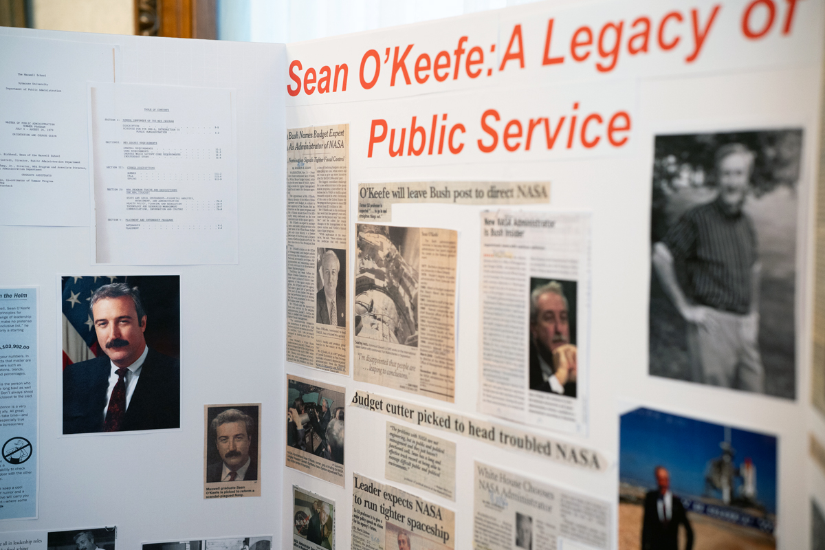 A display board titled 'Sean O'Keefe: A Legacy of Public Service' features newspaper clippings, photographs, and documents highlighting Sean O'Keefe's career. Headlines include 'O'Keefe will leave Bush post to direct NASA,' 'Budget cutter picked to head troubled NASA,' and 'Leader expects NASA to run tighter spaceship.' Several photos of O'Keefe and others are included.