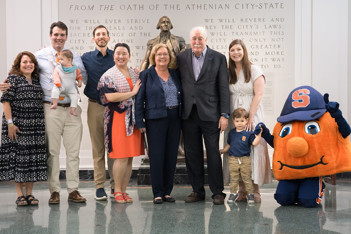 A group of nine people, including adults and children, pose for a photo in front of a statue and an inscribed wall. The inscription is a civic pledge from the Oath of the Athenian City-State. On the right side of the group stands a person in an orange mascot costume with a blue cap featuring an 'S' emblem.