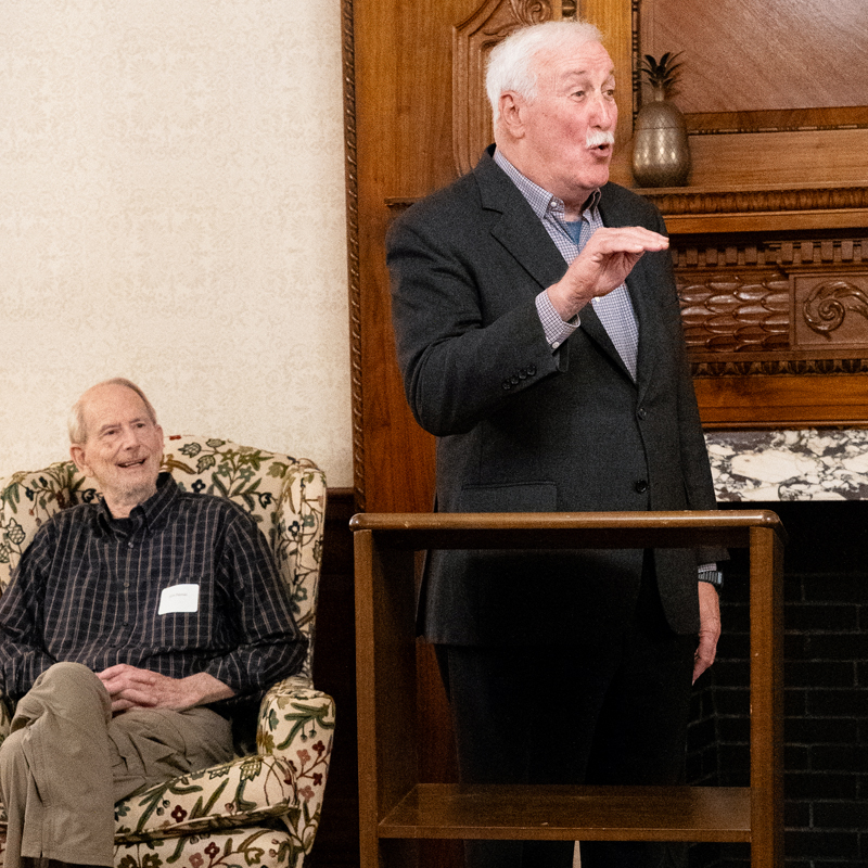 Two men are present in a room with an ornate wooden fireplace and a marble mantlepiece. One man sits on a patterned armchair, wearing a dark checkered shirt and khaki pants with his legs crossed. The other man stands behind a wooden podium, dressed in a dark suit jacket and light blue shirt.
