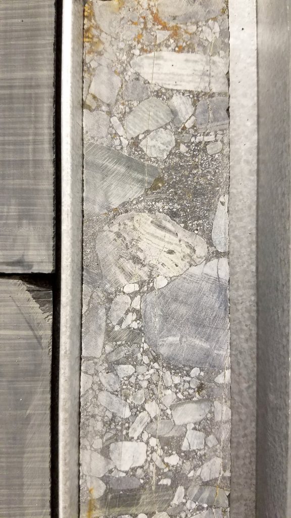 Close-up view of a concrete core sample encased in a metal frame, showing a heterogeneous mix of aggregate materials including small pebbles, larger stones, and fine particles embedded in a cement matrix. The texture and composition of the concrete are clearly visible.