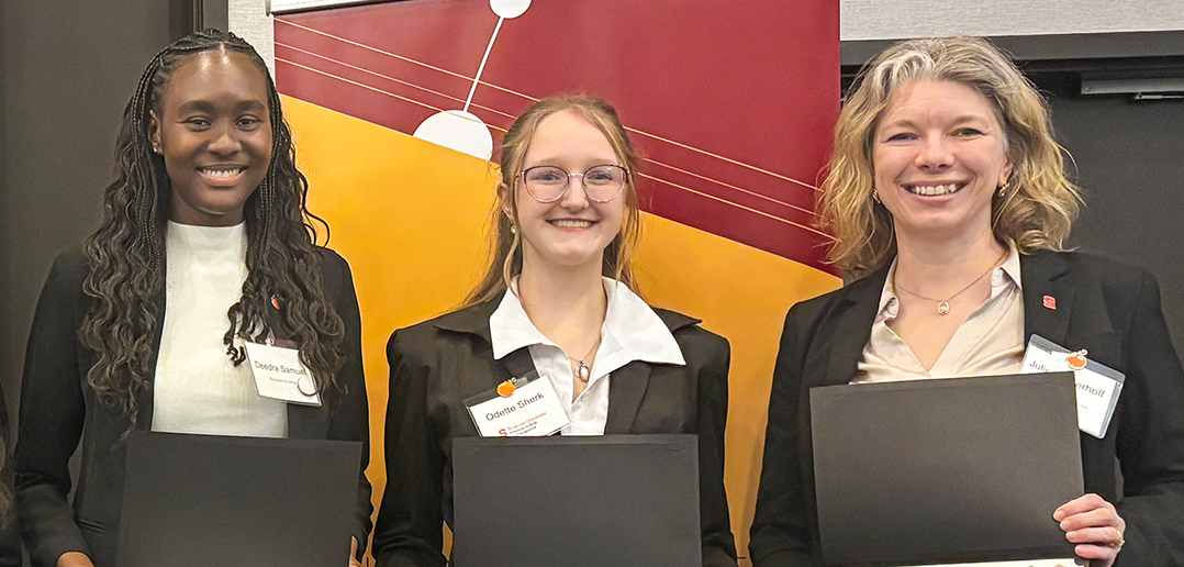 Three individuals stand side by side, each holding a black folder and wearing formal attire with visible name tags. The background features a geometric design in red, yellow, and white. 