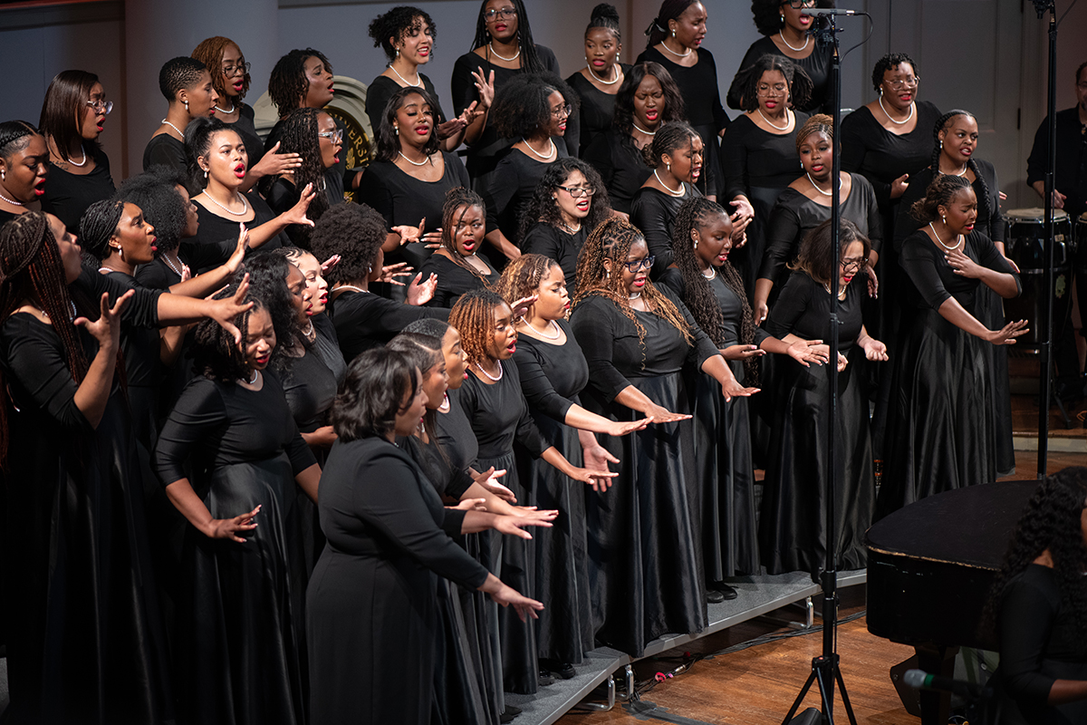 A large choir stands on tiered risers, all dressed in long black dresses. The choir members are mid-performance, with some gesturing expressively with their hands. A microphone stand is positioned in front of the group, and a piano is partially visible in the bottom right corner of the image.