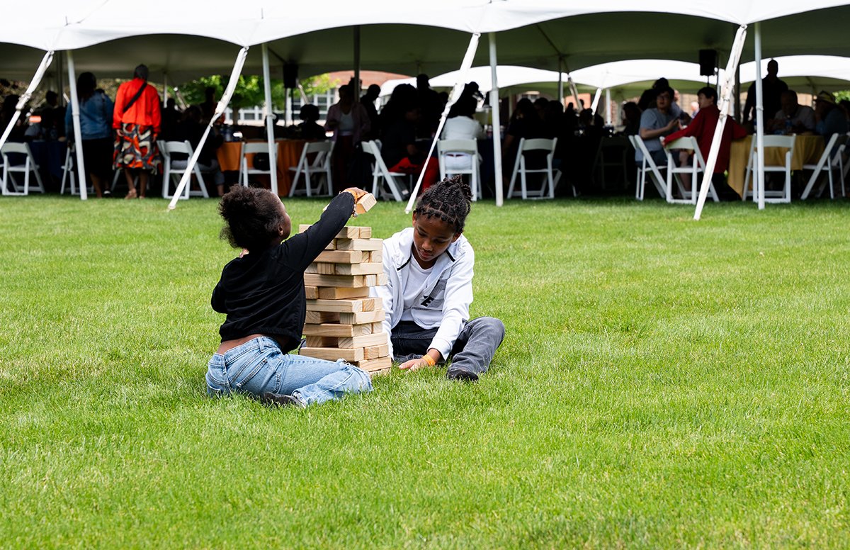 Two children sit on the grass playing a game of giant Jenga. The child on the left wears a black top and blue jeans, and the child on the right wears a white shirt and dark pants. They are focused on stacking large wooden blocks. In the background, people are seated under large white tents.