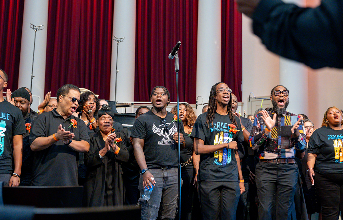 A group of people stands on a stage in front of red curtains and white columns. They are dressed in black clothing, some with colorful designs or text, and are part of a community choir. A microphone stand is positioned at the center. Some individuals are clapping, while others hold water bottles or have their hands at their sides.