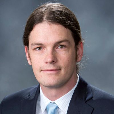 A person with long hair tied back, wearing a dark suit jacket, white shirt, and light blue tie. The background is a plain gray gradient.
