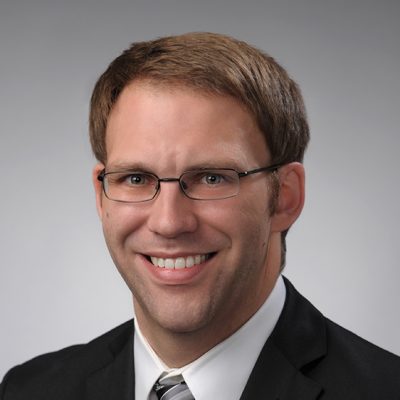 A professional headshot of a person with short brown hair, wearing a black suit jacket, white dress shirt, and striped tie. The background is a plain gray gradient.