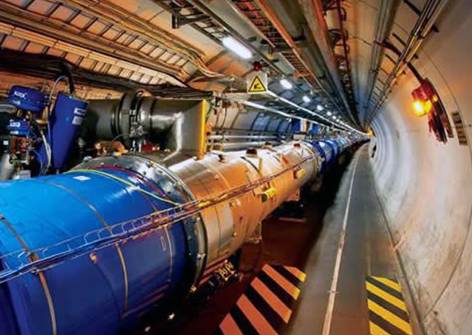 A section of the Large Hadron Collider (LHC) tunnel at CERN, featuring a long, segmented metallic structure with blue and silver components used in particle acceleration. The cylindrical tunnel is lined with cables, pipes, and machinery, illuminated by overhead lights. Safety markings in black and yellow stripes are visible on the floor, along with warning signs along the tunnel walls.