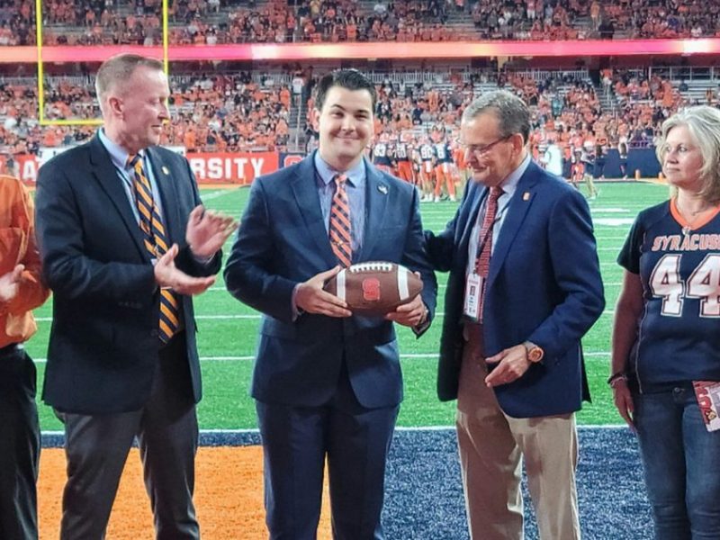 A ceremonial presentation taking place on a football field at Syracuse University. Five people are standing on the field near the end zone. In the center of the group, a man in a dark suit is holding a football, appearing to be receiving it as part of a ceremony. To his left, another man in a suit with an orange and blue striped tie is applauding. On the far left, an older man in an orange collared shirt is also clapping. To the right of the man with the football, an older gentleman in a dark suit jacket appears to be part of the presentation. On the far right, a woman wearing a dark Syracuse jersey with the number 44 is observing the ceremony.