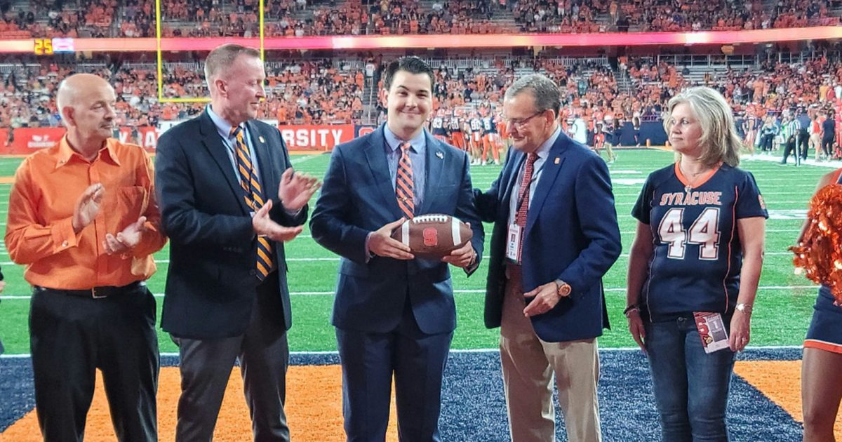 This image shows a ceremonial presentation taking place on a football field at Syracuse University. Five people are standing on the field near the end zone. In the center of the group, a man in a dark suit is holding a football, appearing to be receiving it as part of a ceremony. To his left, another man in a suit with an orange and blue striped tie is applauding. On the far left, an older man in an orange collared shirt is also clapping. To the right of the man with the football, an older gentleman in a dark suit jacket appears to be part of the presentation. On the far right, a woman wearing a dark Syracuse jersey with the number 44 is observing the ceremony.