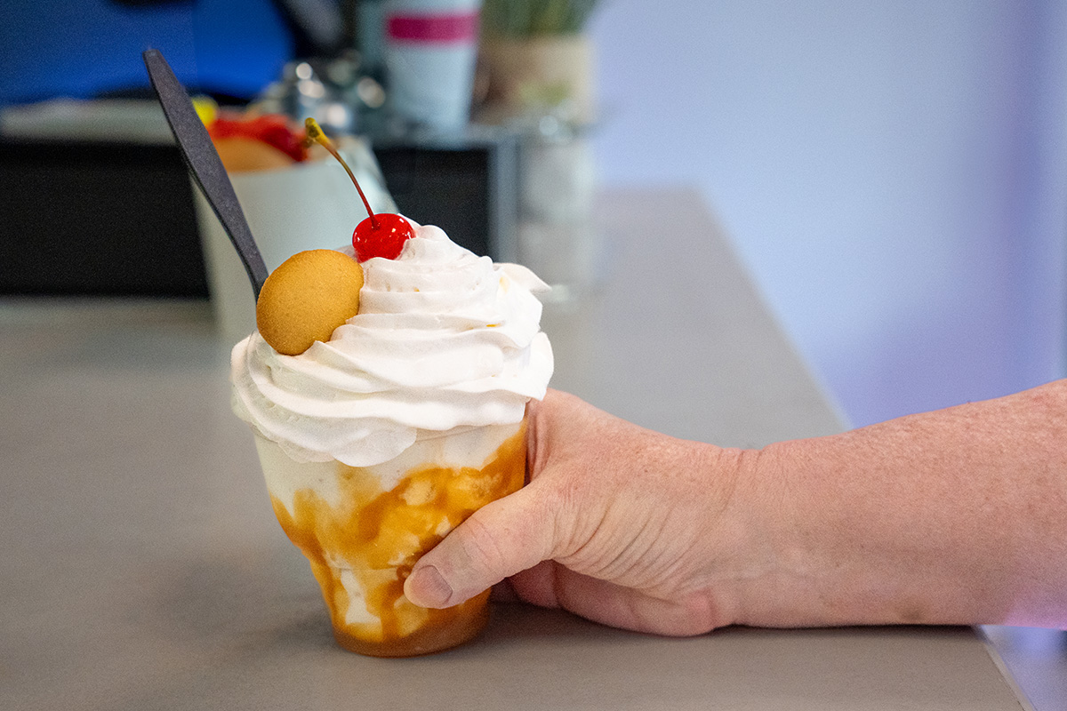Hand holding a dessert cup filled with ice cream, topped with whipped cream, a cherry, caramel drizzle, and a vanilla wafer cookie, with a black plastic spoon inserted; blurred indoor background with counter items.