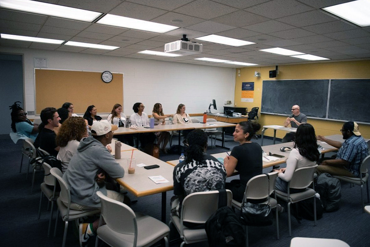 A classroom with students seated around a large U-shaped table, facing an instructor at the front. The room features fluorescent lighting, a ceiling-mounted projector, and a large chalkboard behind the instructor. Tables are scattered with notebooks, water bottles, and drinks. Posters decorate the walls, and a clock hangs above a bulletin board.