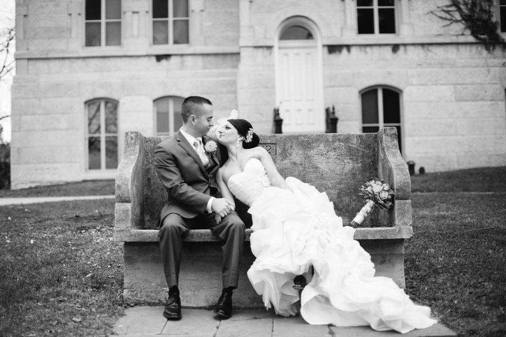 A black and white photograph of a couple seated on a stone bench in front of a historic building with arched windows and a large door. The person on the left wears a formal suit, and the person on the right wears a wedding dress and holds a bouquet. The setting includes a grassy area and the textured stone facade of the building.