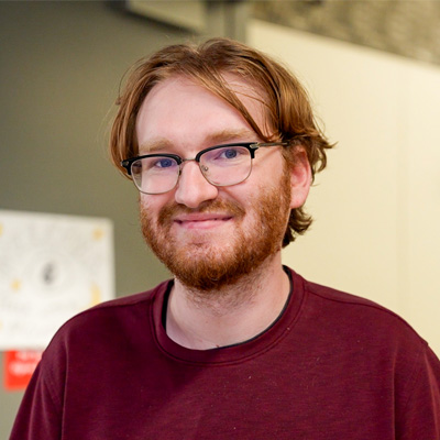 Person with medium-length reddish-brown hair and beard wearing a maroon shirt, standing in front of a blurred wall.