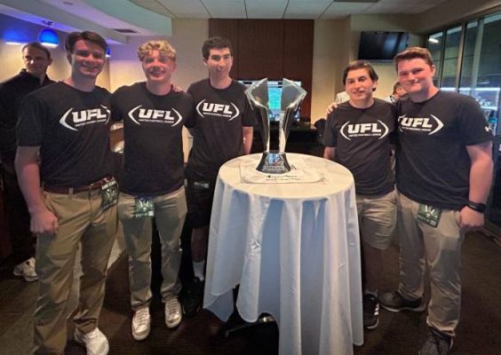 Five young men in black UFL t-shirts stand around a table